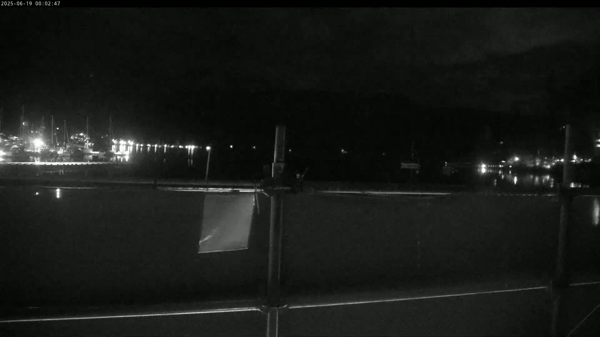 A nighttime view of a calm harbor with boats docked, seen from behind a metal fence.