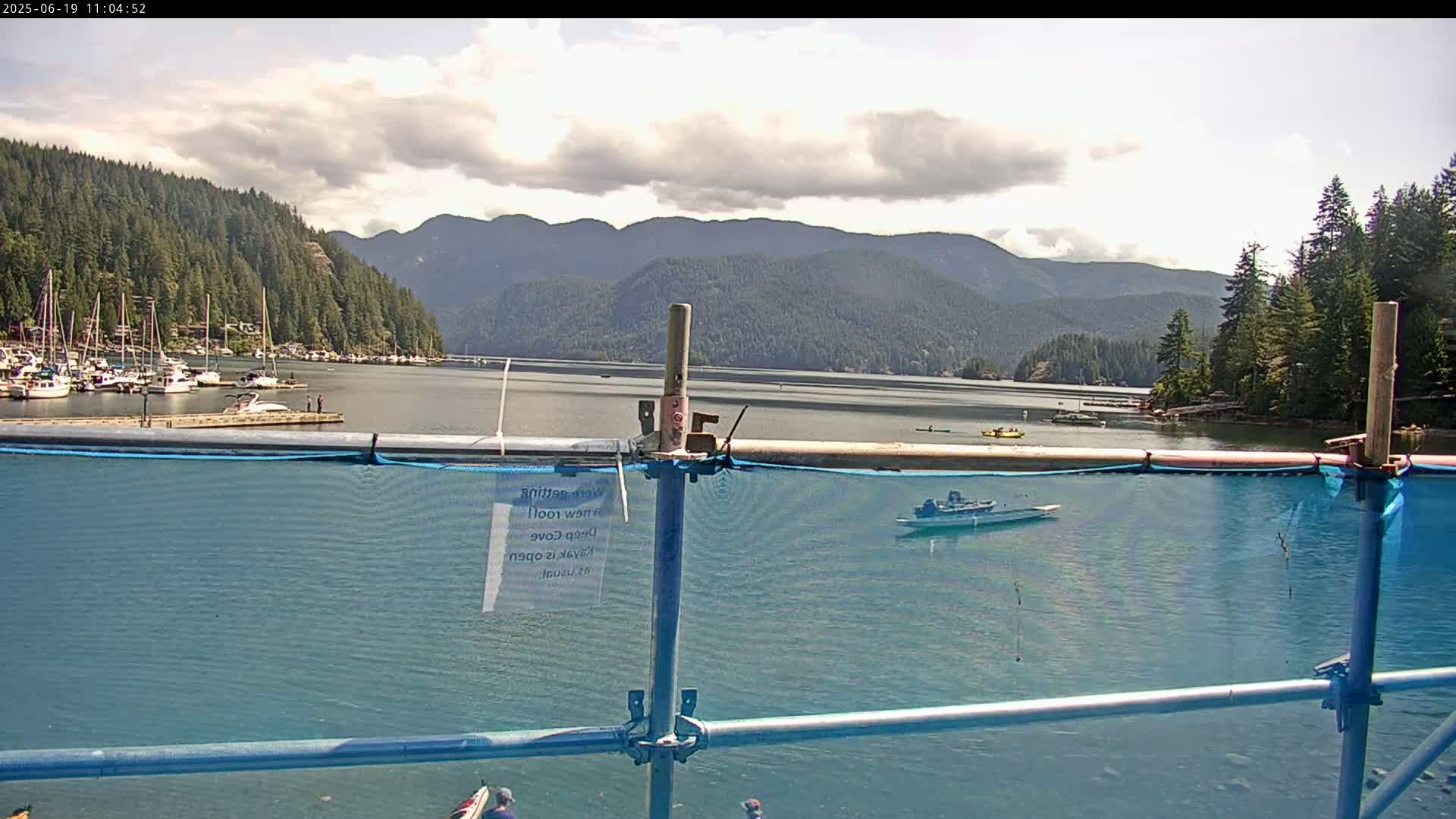 A partly cloudy day shows a calm bay with boats moored at a dock, a few small boats on the water, and mountains covered in evergreen trees in the background, all viewed through a blue mesh safety fence.
