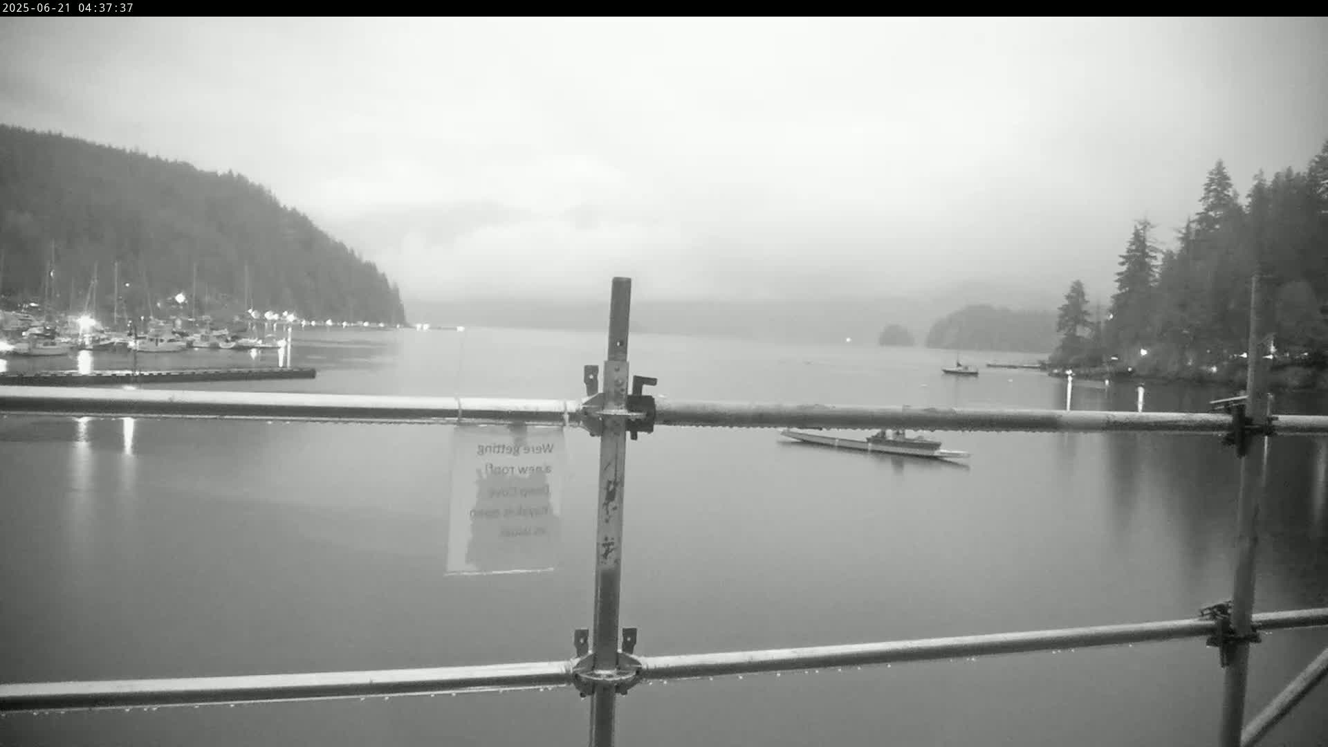A calm body of water with boats and a shoreline lined with trees is viewed through a metal railing in the rain.