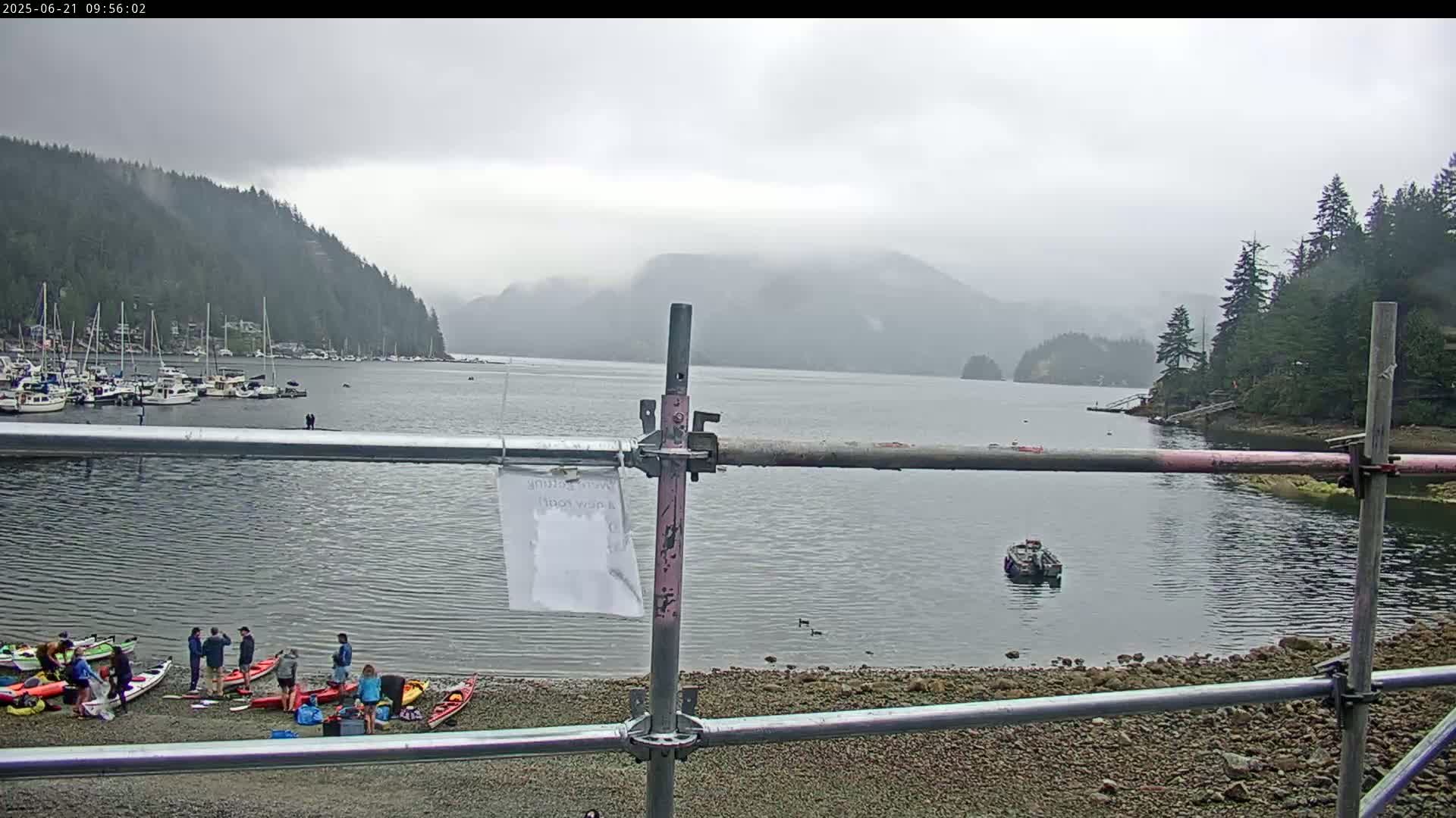 A calm bay with a group of people near kayaks on a rocky shore, under an overcast sky, is partially obscured by a metal scaffolding.