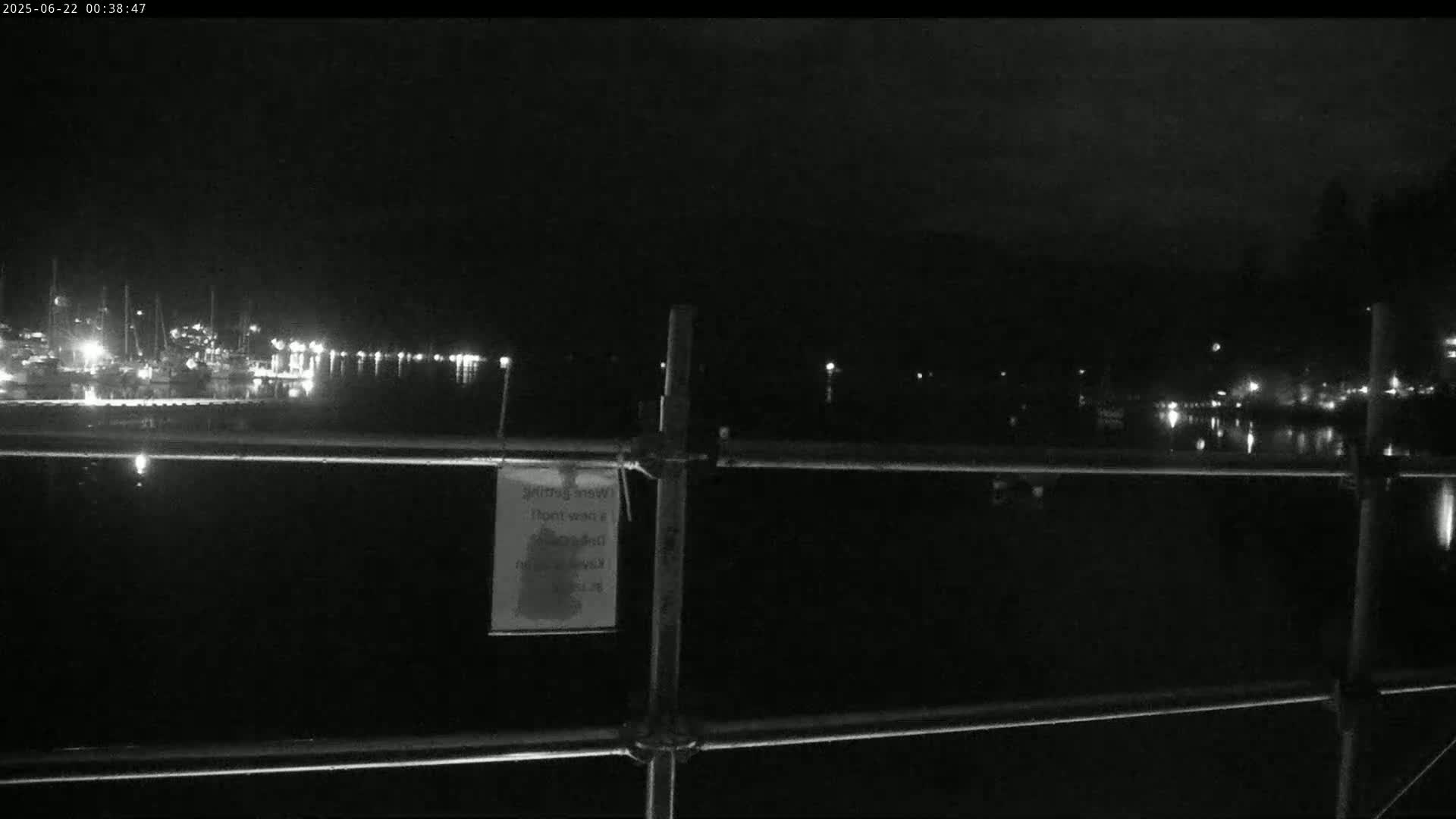 A nighttime view of a calm body of water with a marina and lights in the distance, seen from behind a metal fence.