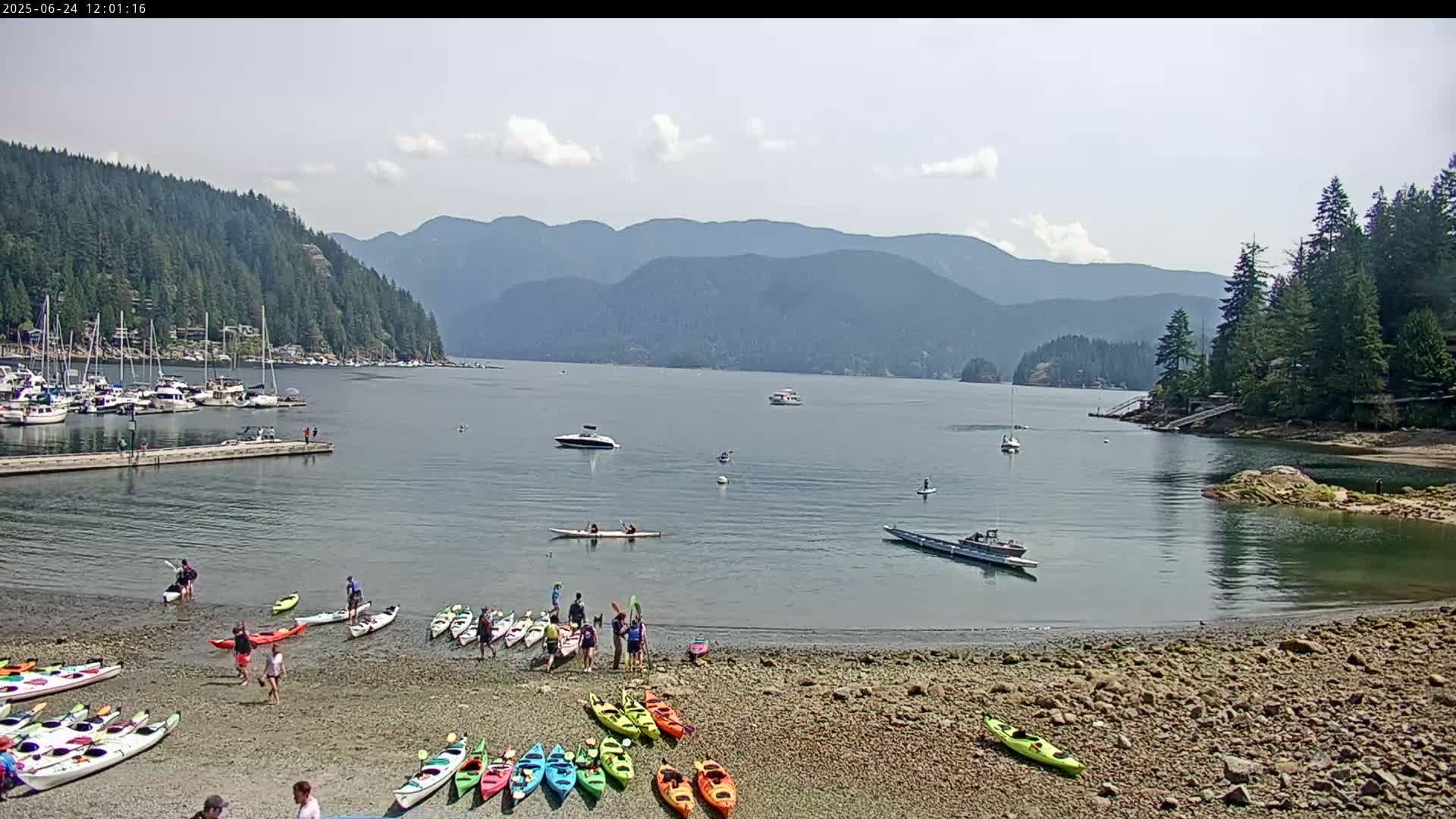 A sunny day on a rocky beach with numerous kayaks and people, boats on the water, and forested mountains in the background.