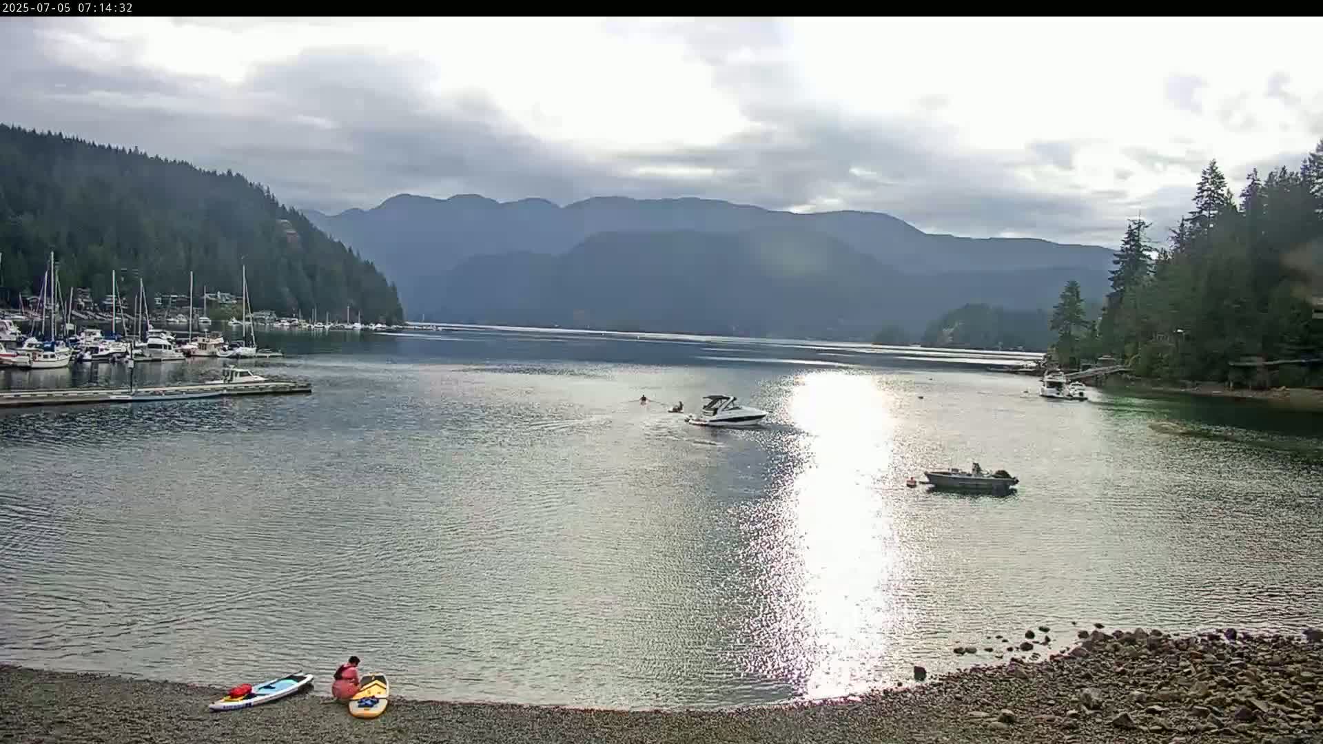A calm bay with several boats and two people preparing paddleboards under a mostly cloudy sky, backed by forested mountains.