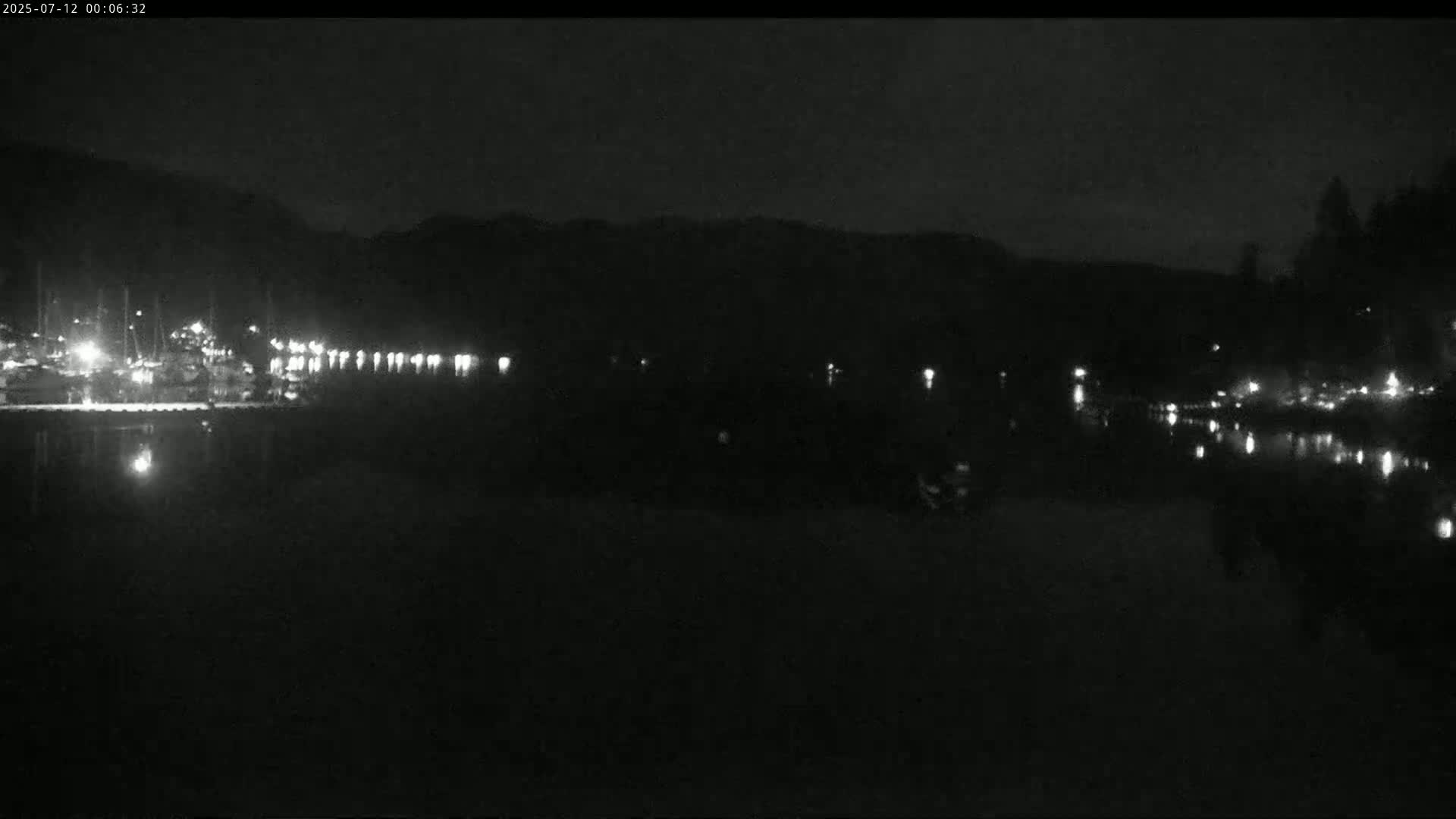 A nighttime view of a calm body of water reflecting the lights of a marina and shoreline, under a dark sky.