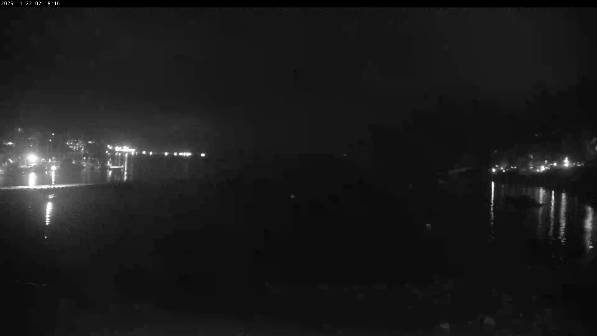 A dark night scene captures a wide body of water calmly reflecting numerous bright lights from distant structures along its left and right banks.