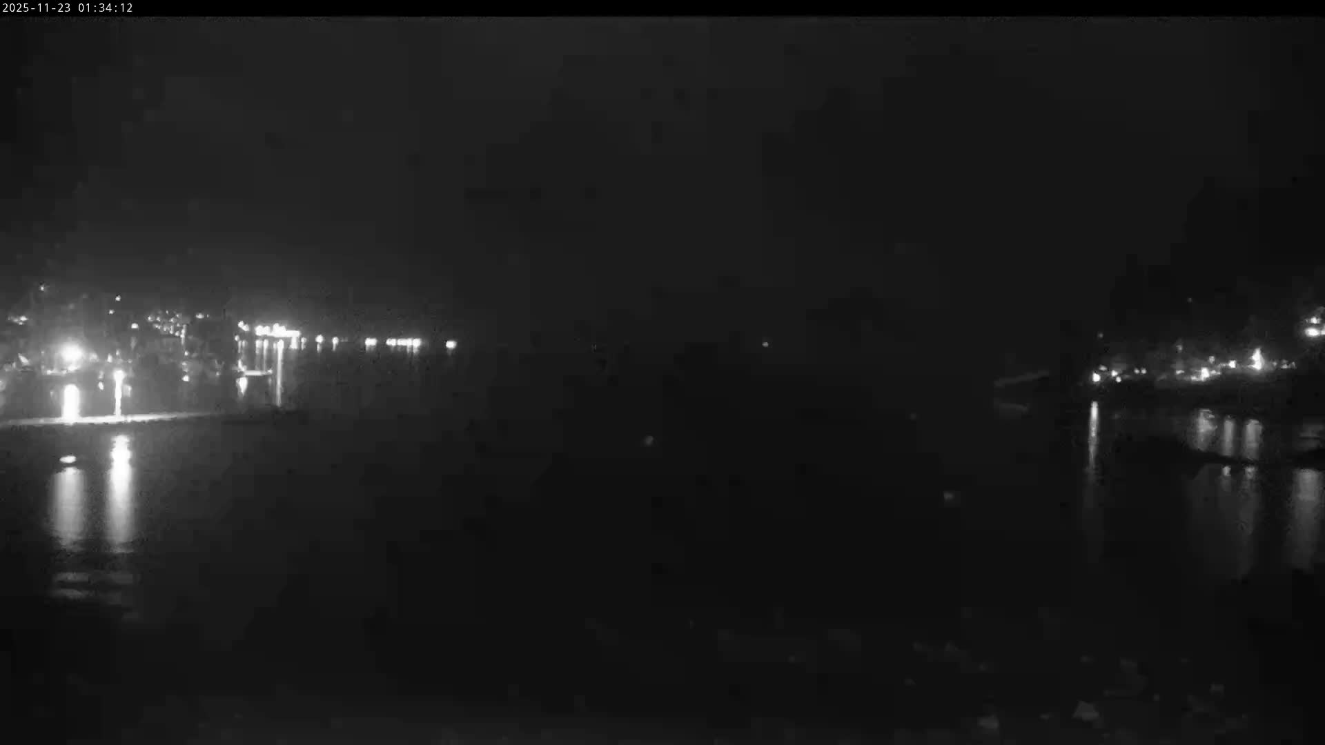 The image displays a very dark and foggy night scene over a body of water, with faint city lights reflecting on the water from both the left and right distant shores.