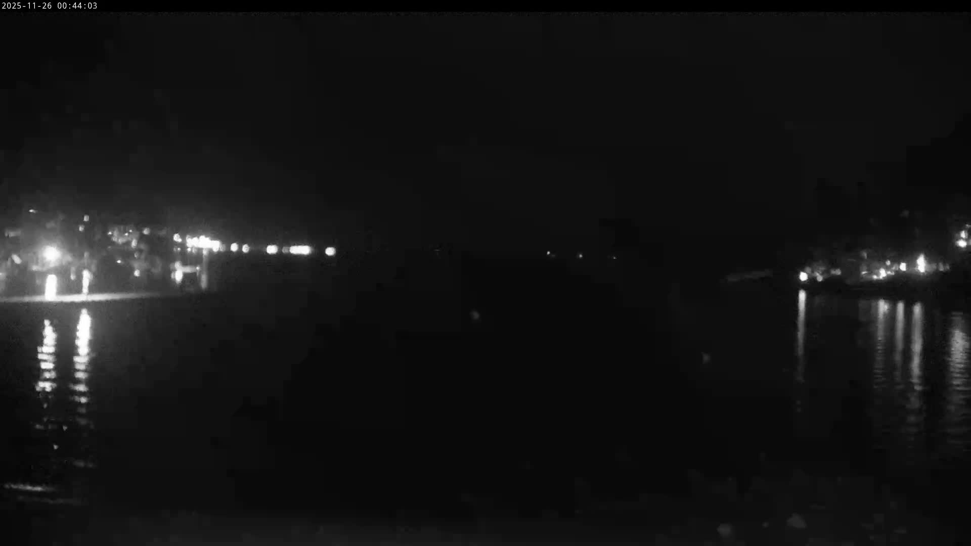 A dark night scene displays numerous distant lights reflecting on a calm body of water, indicating clear weather.