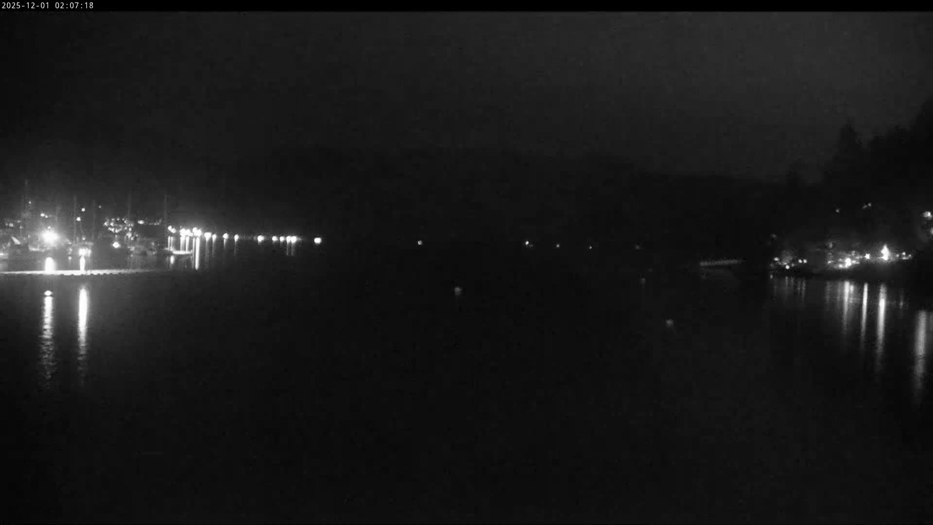 A dark, clear night shows a calm body of water reflecting numerous lights from a distant shoreline, with a marina visible on the left and illuminated structures on the right.