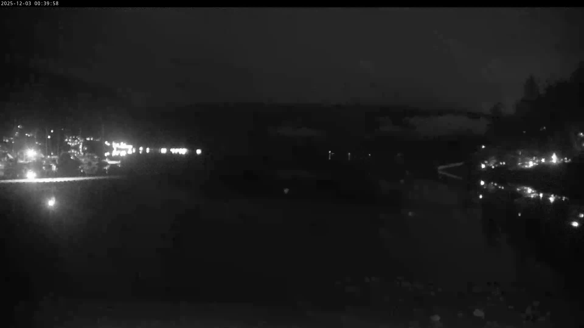 It is a dark, cloudy night overlooking a body of water, with numerous lights from settlements or structures illuminating both sides and reflecting in the water.