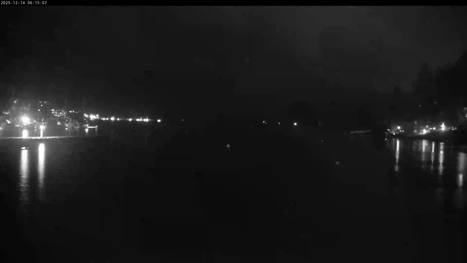 It is a dark, cloudy night overlooking a body of water, with numerous lights from settlements or structures illuminating both sides and reflecting in the water.