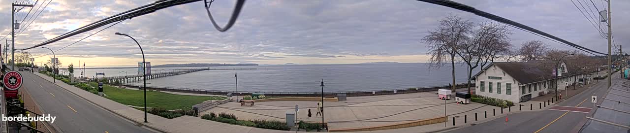 A panoramic view of a waterfront town on an overcast day shows a long pier extending into a calm sea, with buildings, trees, and a paved area situated along the shore.