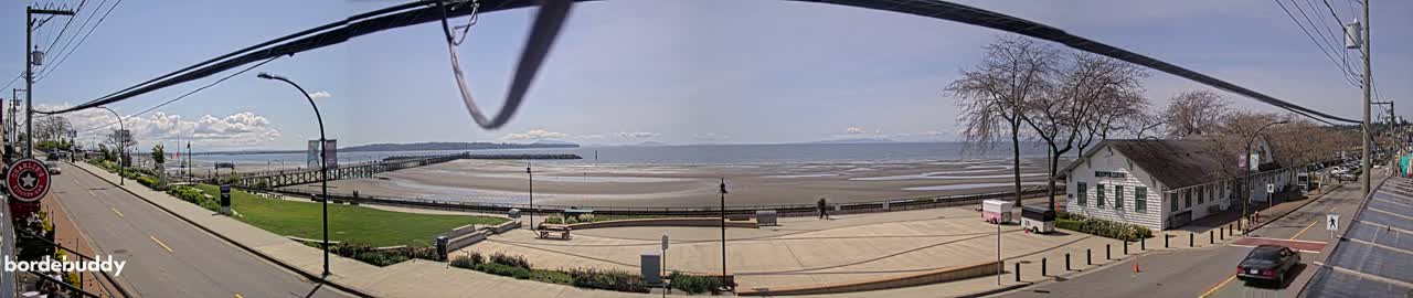 A panoramic view shows a partly sunny day overlooking a beach with a pier, a street with parked cars, and a building near the water's edge.