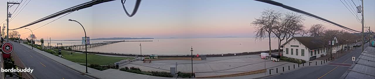 A waterfront scene at dawn shows a long pier extending into calm water, a paved area with a building and trees, and a street with power lines overhead.