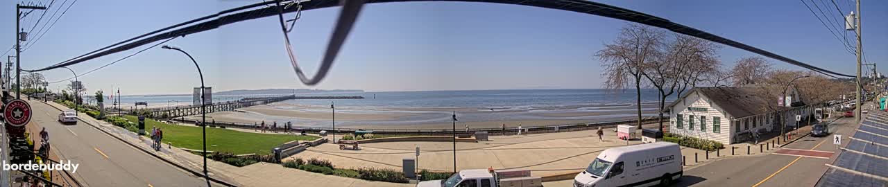 A sunny day overlooking a beach with a pier, a grassy area, buildings, and vehicles parked along a street.