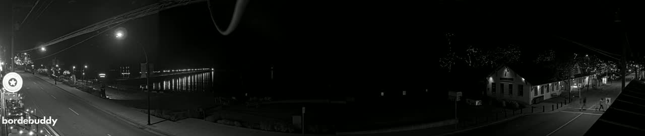A nighttime, black and white view of a town street leading to a lit pier extending into the water, with a building visible on the right.