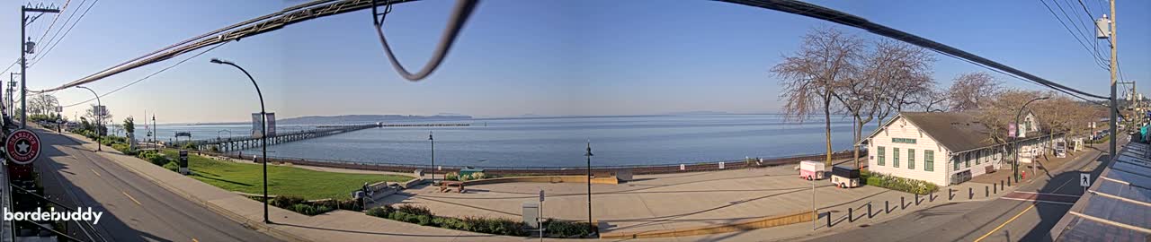 A panoramic view of a waterfront scene on a sunny day shows a long pier extending into a calm body of water, a grassy park area, a white building, and a street with overhead power lines.