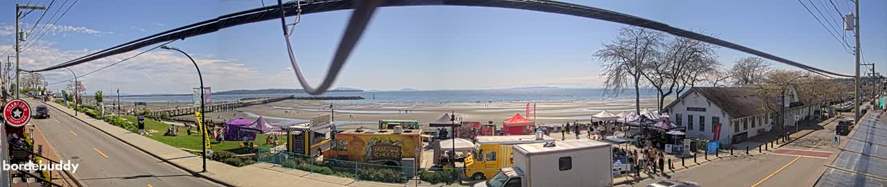 A sunny beach scene with food trucks and market stalls, situated beside a road and a row of buildings, all under a clear blue sky.