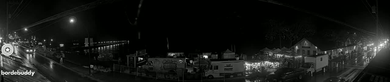 A nighttime, rain-slicked street scene shows a collection of food trucks and market stalls, illuminated by lights, alongside a waterfront with buildings and trees in the background.