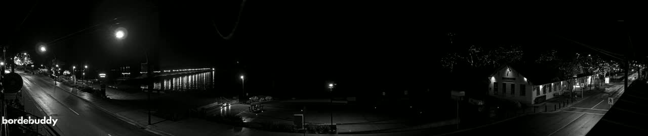 A nighttime black and white panoramic view shows a wet, empty street lined with buildings and lights, a pier extending into the water, and a building with lights in the distance.