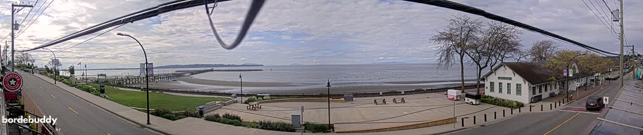 A panoramic view of a beach town on a partly cloudy day shows a calm body of water, a long pier, a grassy park, and a street with buildings.