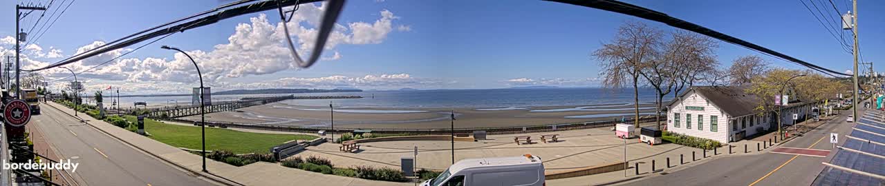 A sunny day reveals a waterfront scene with a sandy beach, a pier, a white building, and a street with parked vehicles, under a partly cloudy sky.