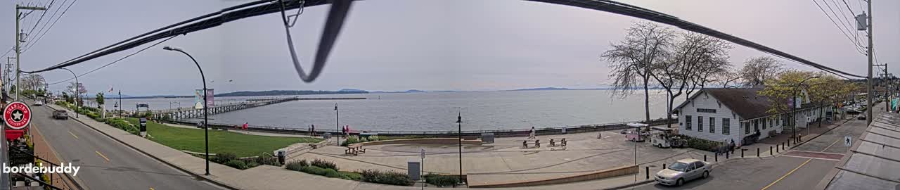 A waterfront scene on a partly cloudy day shows a pier extending into calm water, a paved plaza with picnic tables, a white building, and a street with cars.
