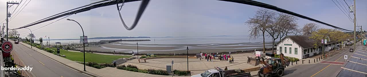 A sunny day reveals a seaside town with a long pier, a sandy beach at low tide, a group of people gathered in a plaza, and a road lined with buildings, including a white structure that appears to be a historical building.