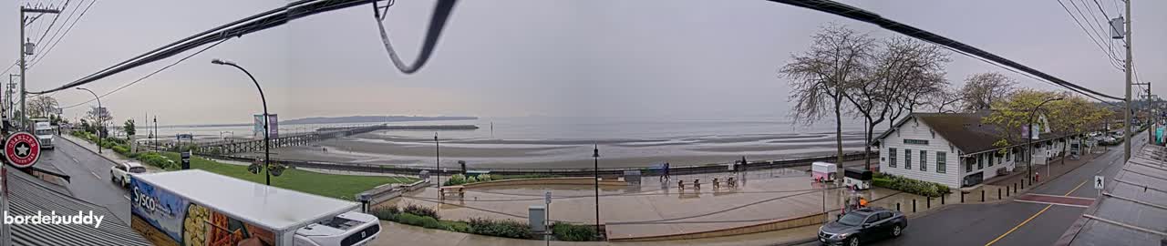 A seaside town scene on an overcast day shows a wet paved area near the ocean, a long pier extending into the water, and a street with cars parked along a row of buildings.