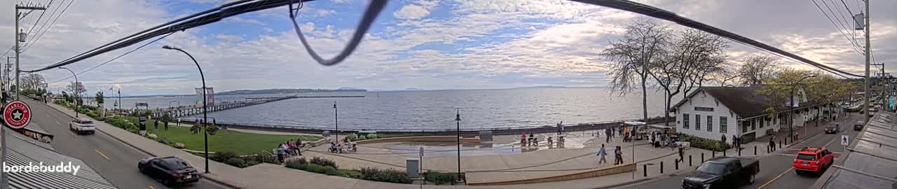 A waterfront scene on a partly cloudy day shows a pier extending into a calm body of water, with people gathered in a plaza near a building and cars parked along a street.