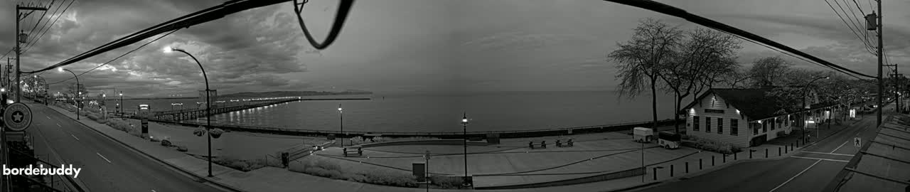 A grayscale panoramic view shows a waterfront scene at dusk under cloudy skies, featuring a street, a pier extending into the water, and a building near the shore.