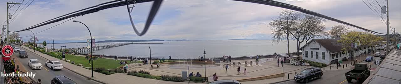 A partly cloudy day overlooks a waterfront scene with a long pier, a plaza with people, and cars parked along a street next to buildings.
