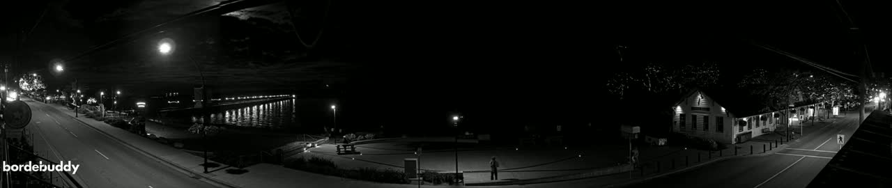 A nighttime, black-and-white panorama shows a street scene with a waterfront, buildings, and a few people, under a partly cloudy sky.