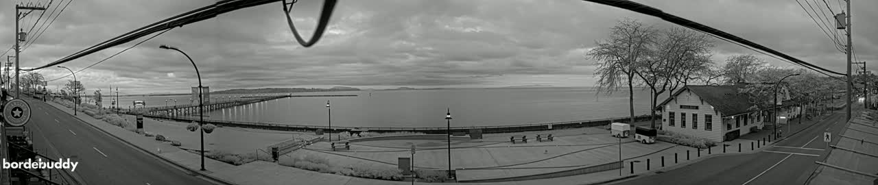 A grayscale panoramic view shows a waterfront scene with a pier, a road, and buildings under a cloudy sky.