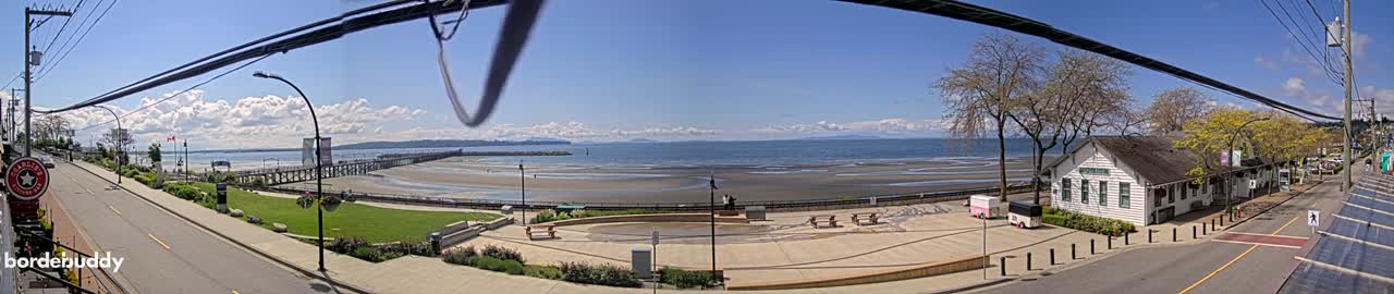 A panoramic view of a waterfront town on a sunny day, showing a sandy beach, a pier, a small plaza, and buildings, with a calm ocean extending to distant mountains under a partly cloudy sky.