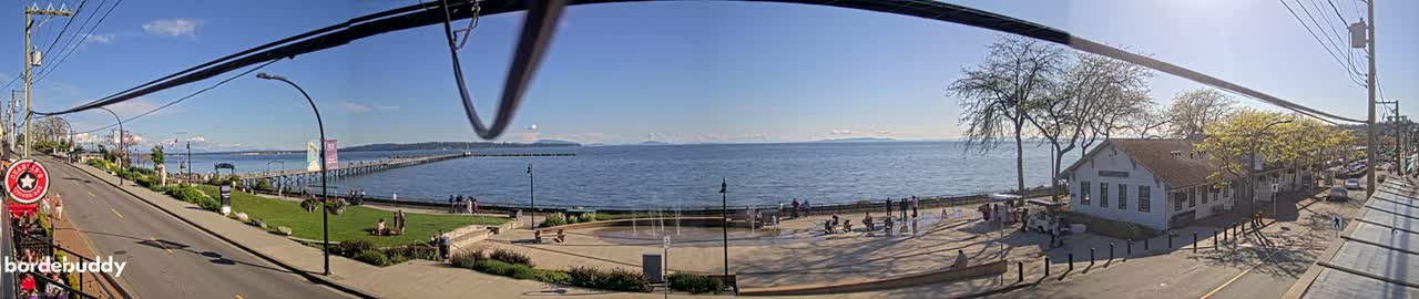 A sunny waterfront scene shows a pier extending into a calm bay, a town with buildings and a park with a water feature, all under a clear blue sky.