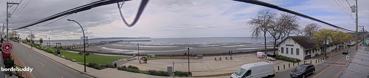 A panoramic view of a seaside town on an overcast day, showing a beach, a pier, a street with cars, and buildings, including a white building near the water.