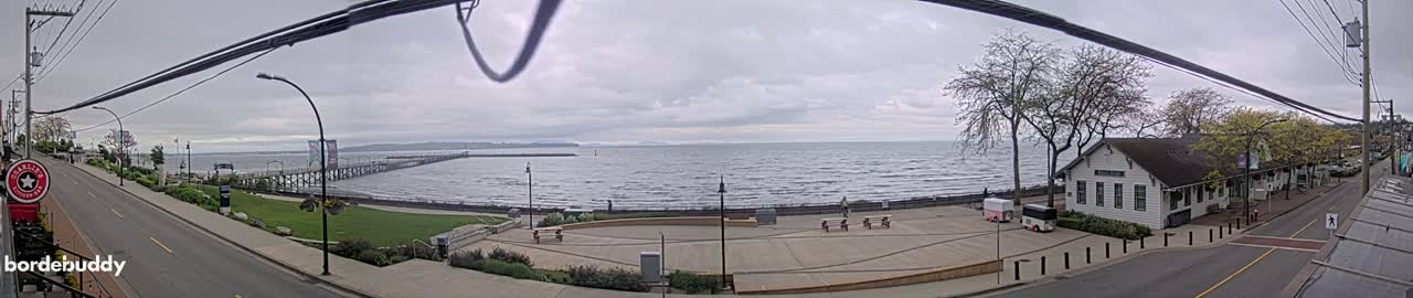 A waterfront scene on an overcast day shows a long pier extending into a choppy sea, with a paved area, buildings, and trees along the shore.