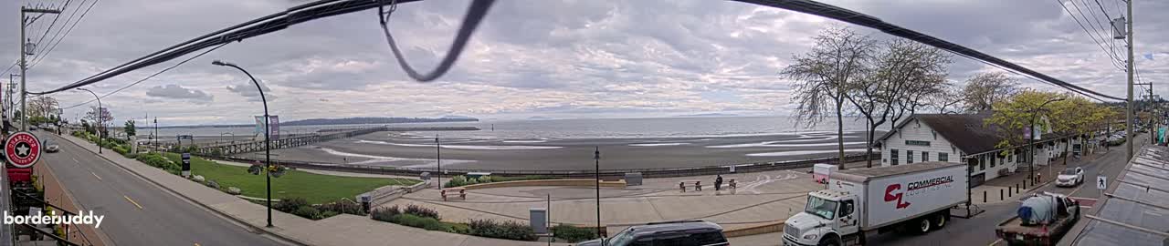 A panoramic view shows a mostly cloudy sky over a beach and ocean, with a town’s buildings and streets visible in the foreground.