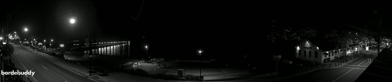 A black and white nighttime panorama shows a sparsely populated street curving past buildings and a pier with lights reflecting on the water.