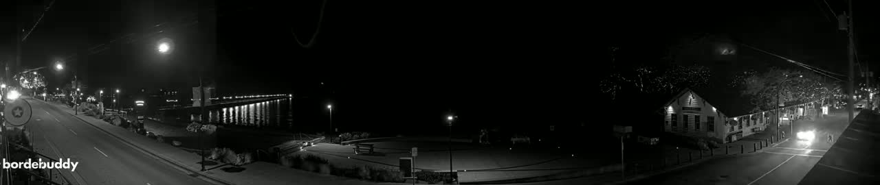 A nighttime black and white view of a street scene with a waterfront pier, buildings, and a car driving down the road.