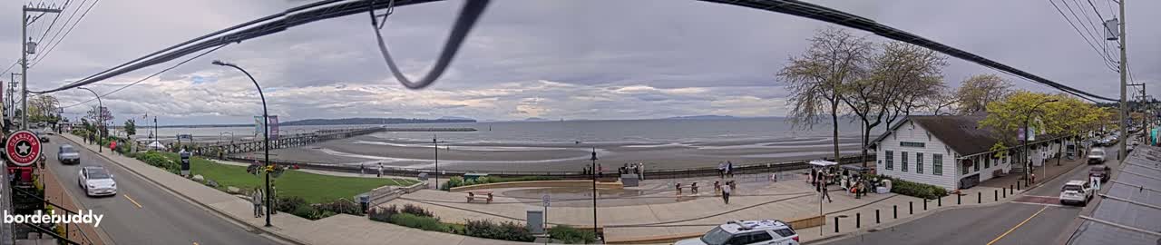 A panoramic view of a seaside town on an overcast day shows a beach, a pier, a waterfront plaza with people, and a street with cars parked alongside buildings.