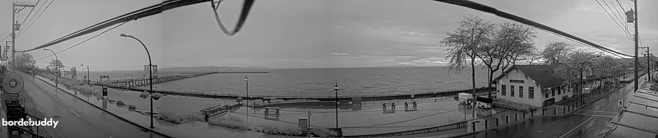 A black and white, wide-angle view shows a waterfront scene on a cloudy day with flooded areas near a pier and buildings, and a road with power lines overhead.