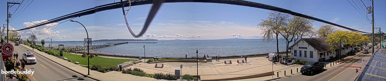 A sunny day overlooks a waterfront town with a pier, a paved plaza, and a calm ocean with distant mountains under a partly cloudy sky.