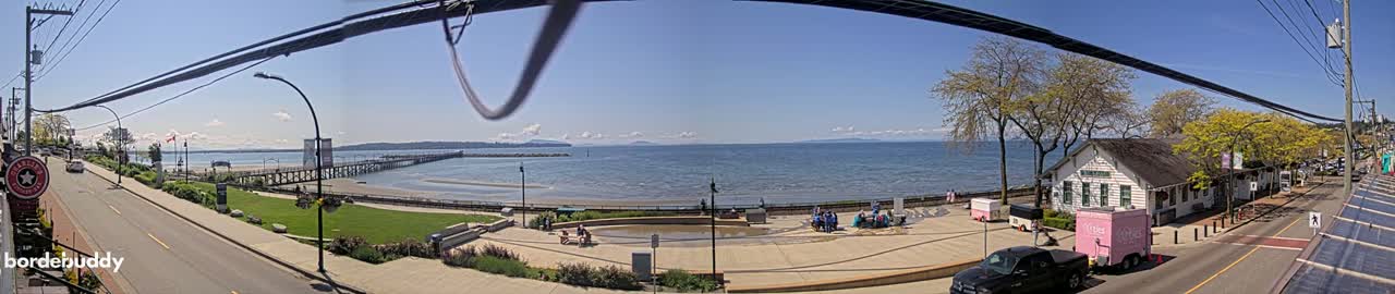 A seaside town scene on a sunny day shows a paved waterfront area with people gathered near food trucks, a pier extending into calm water, and a street with cars along a tree-lined roadway.
