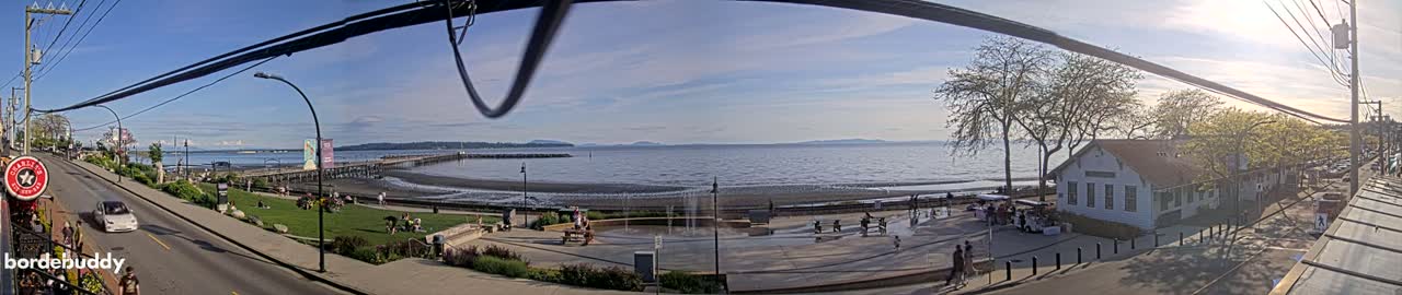 A seaside town on a sunny day, with a long pier extending into calm water, shows people relaxing on the beach and in a plaza near a white building, all under a clear blue sky.