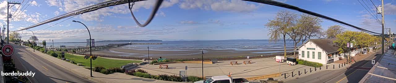 A panoramic view of a seaside town on a partly cloudy day shows a beach, a pier, a waterfront promenade with picnic tables, and buildings along a street.