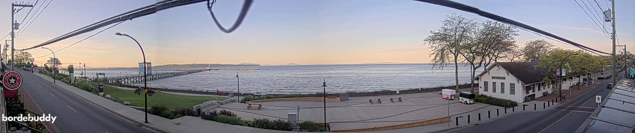 A calm ocean with a long pier extends into it at sunset, next to a paved waterfront area with trees and buildings.