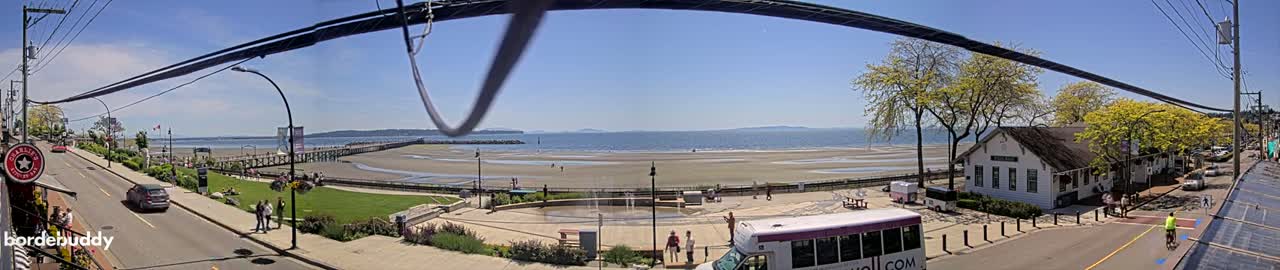 A sunny day reveals a seaside town with a long pier extending into calm waters, a beach, and a street lined with buildings and trees.