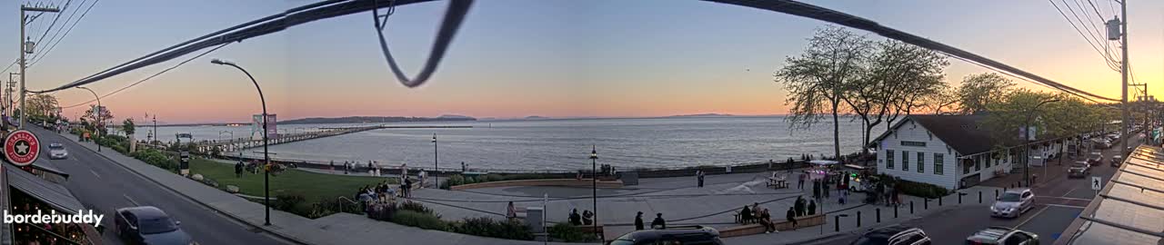 A waterfront scene at sunset shows a calm body of water, a long pier, a town with cars parked along a street, and several people gathered in a plaza area.