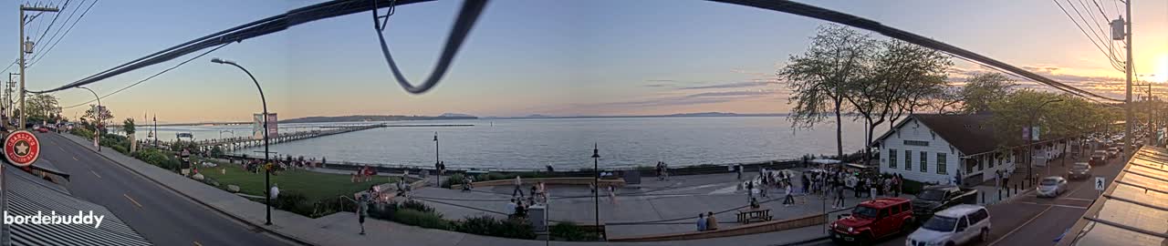 A waterfront scene at sunset shows a calm bay with a pier and a town with people gathered on a plaza near a building, all under a clear sky.